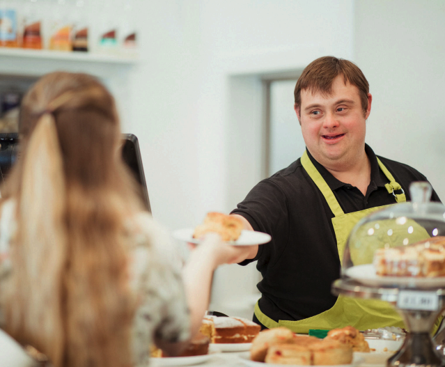 Une jeune homme qui a une trisomie portant un tablier jaune travaille dans un café et sert une viennoiserie à une cliente
