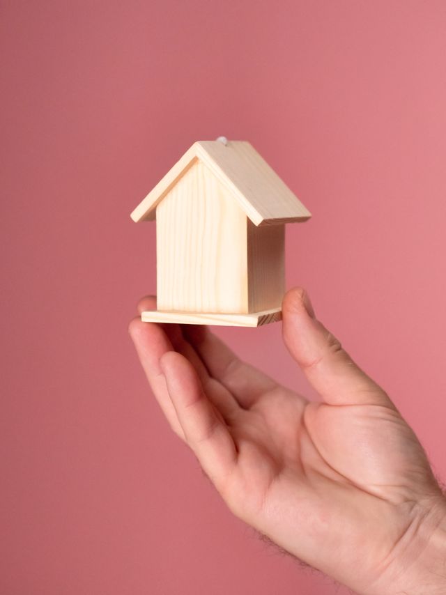 Une main tient une petite maison de bois, sur fond rose.